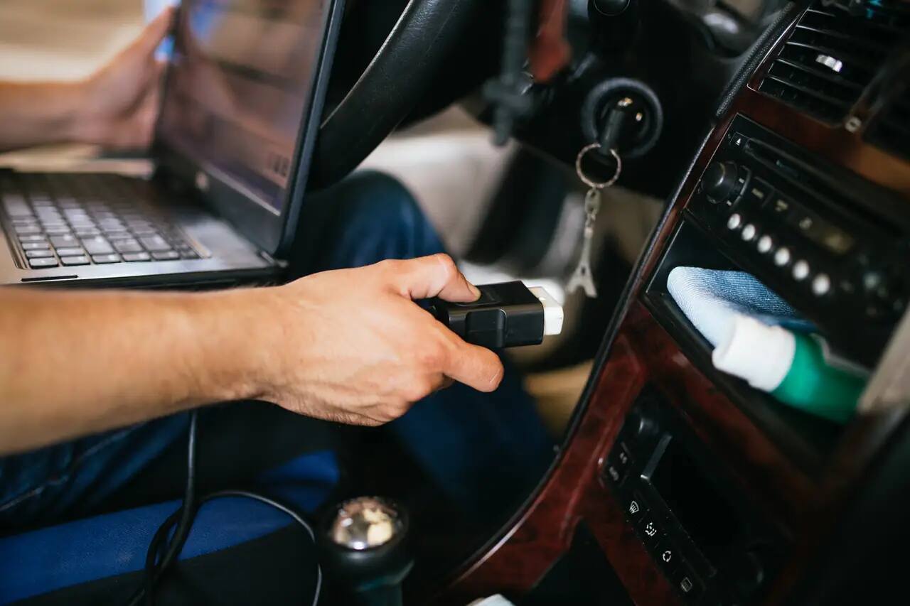 Майстер тримає розʼєм діагностичного адаптера OBD-II під кермом авто, поруч на колінах ноутбук